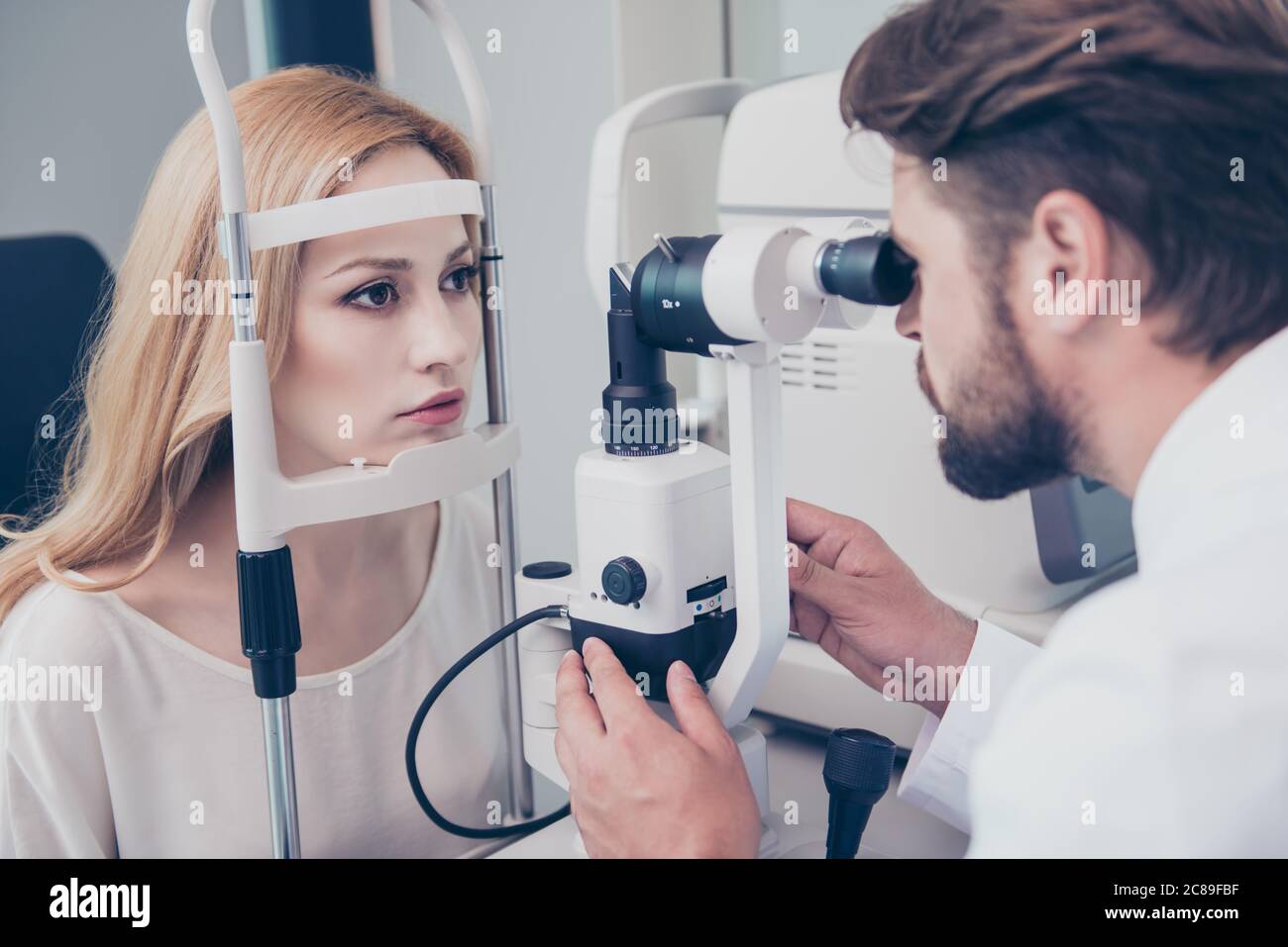 Concentrated brunet bearded optician with non contact tonometer is checking blond`s lady patient intraocular pressure at eye clinic. Health care, medi Stock Photo